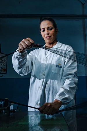 Woman in a white REDA lab coat holds taut dark threads in a textile mill.
