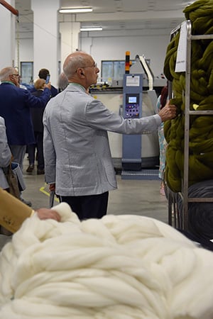 Man in a gray jacket touches a machine in a textile mill, beside large skeins of wool top and a rack of green wool.