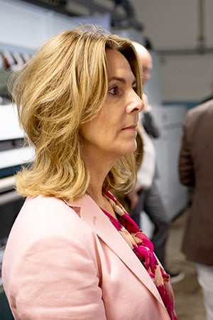 Woman with shoulder-length blonde hair in a pink blazer, in profile, inside a textile production facility