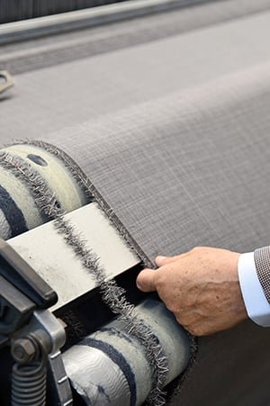 Hand of a man in a suit touching grey woven fabric as it passes over rollers on a textile machine.