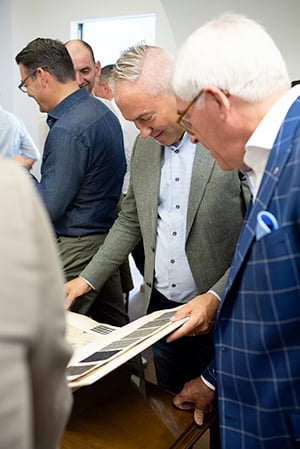 Men in jackets smile as they look through a book of dark fabric swatches on a wooden table.