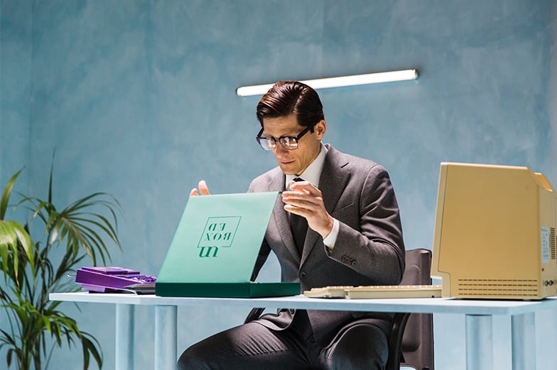Man in a gray suit and glasses opens a green "unboxed" box at a desk with a vintage computer
