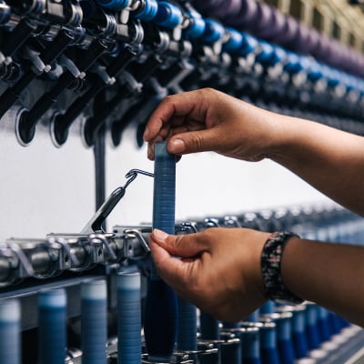 Hands hold a blue yarn bobbin on a textile machine with rows of blue bobbins in the background.