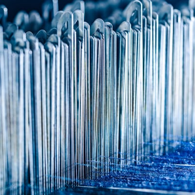 Rows of thin metal heddles on a loom, with blue wool yarn visible below, in blue-toned light