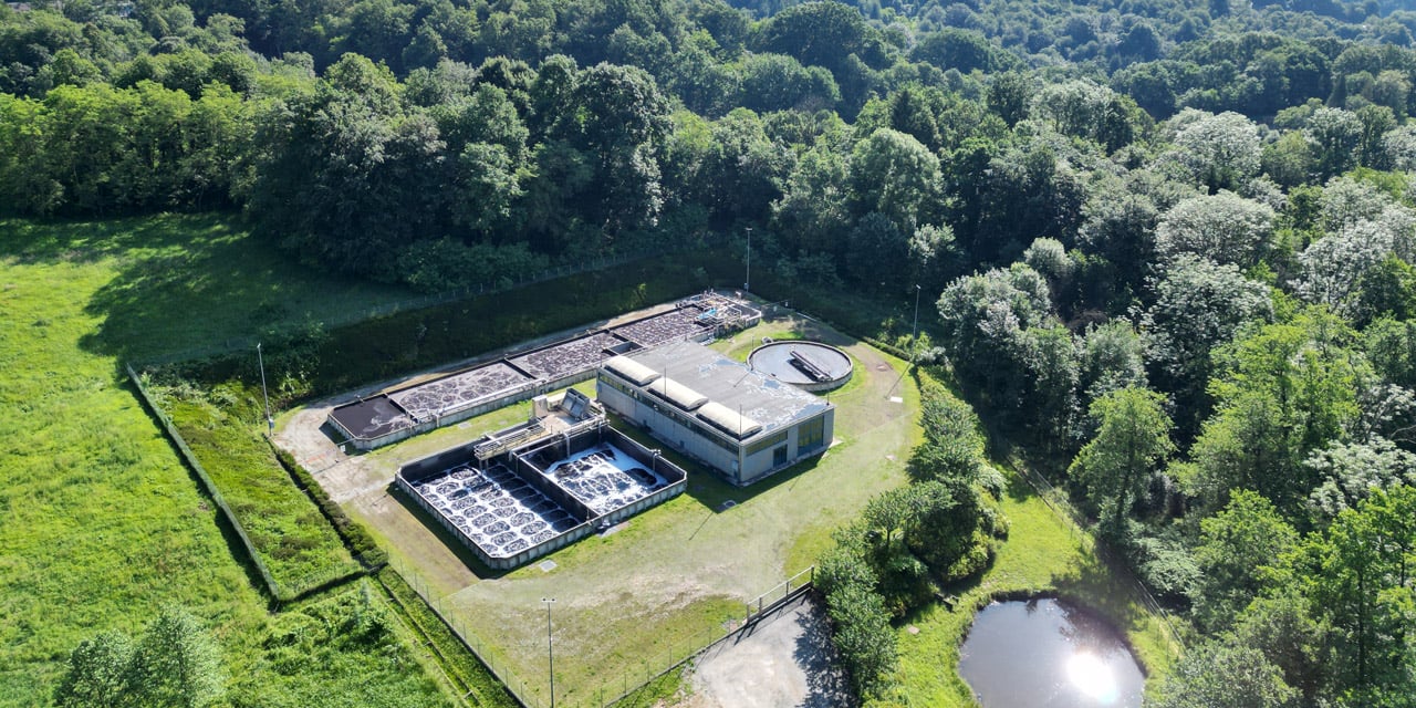 Aerial view of a wastewater treatment plant with circular tanks, surrounded by green meadows and forest