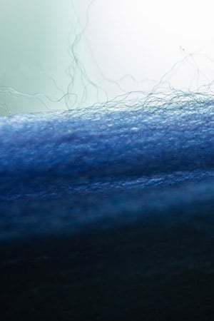 Macro close-up of blue wool fabric with fine loose fibers rising against a soft pale green background