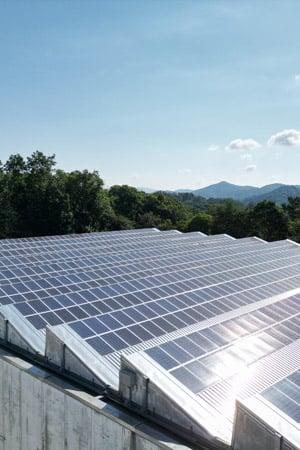 Rows of solar panels on a building roof, with green forested hills and a blue sky in the background