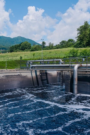 Water basin with foamy dark water and metal pipes at a treatment plant, with green hills and clouds behind