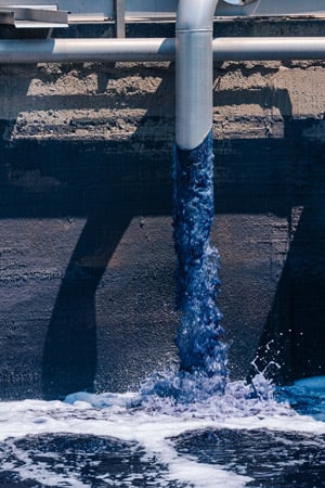 Blue-tinted water pours from a metal pipe into a concrete tank, creating white foam below