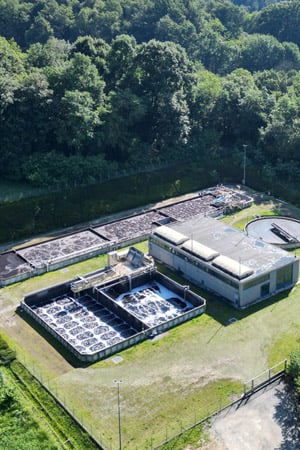 Aerial view of a water treatment plant with open tanks and a building, surrounded by green forest