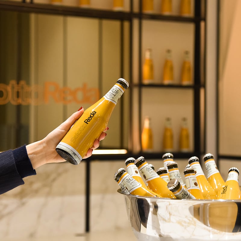 Hand holding a yellow Salotto Reda bottle above an ice bucket of bottles, with shelves behind.