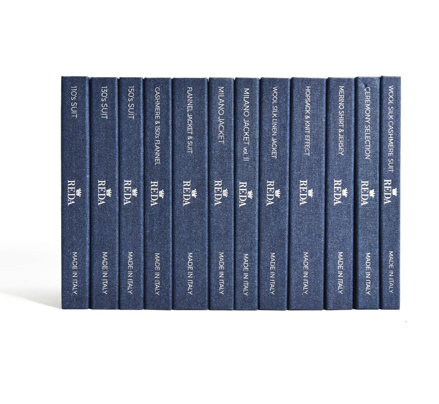 Twelve navy cloth-bound swatch books with REDA logo and "Made in Italy" on the spines, labeled by fabric type