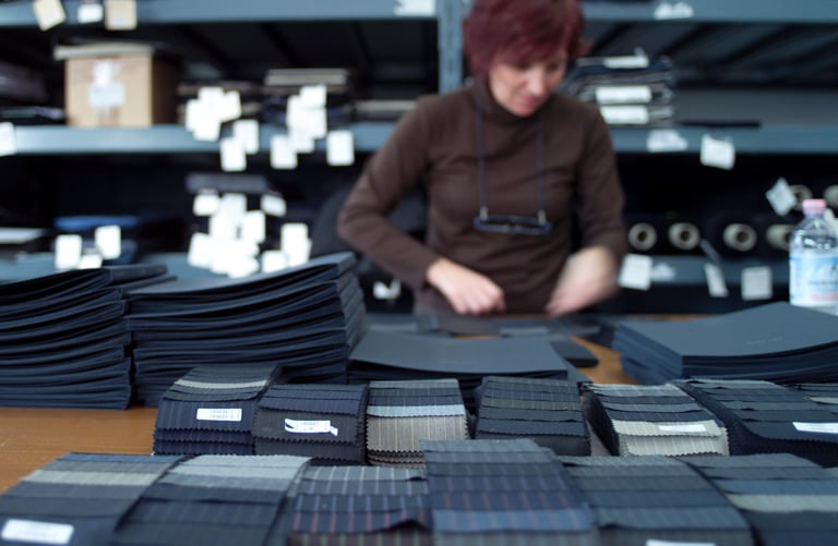 Donna con maglione marrone sistema pile di campioni di tessuto di lana scuro piegati su un tavolo in un magazzino tessile.