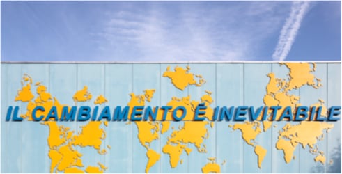 Mappamondo giallo su parete azzurra con la scritta "IL CAMBIAMENTO È INEVITABILE" sotto un cielo blu