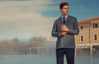 Man in a blue checked double-breasted jacket and tie adjusts his cuff outdoors by a stone building.
