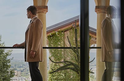 Man in a camel wool coat leans on a balcony railing, looking out over a hilltop landscape.