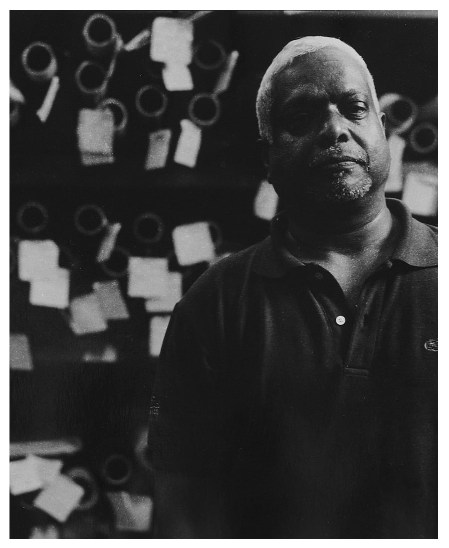 Black-and-white portrait of an older man with white hair in a dark polo shirt, with spools and paper tags behind him