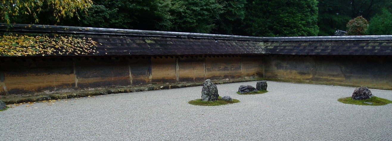Zen rock garden with raked gravel, moss-covered stone groups and a low tiled earthen wall beneath green trees