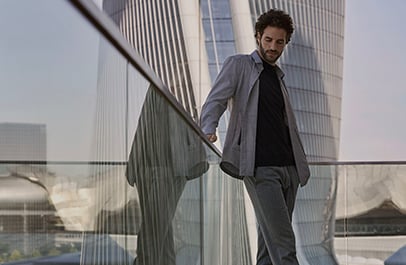 Man in a gray jacket and black shirt leans on a glass railing, with modern skyscrapers behind him.