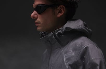 Man in profile wearing dark sunglasses and a grey hooded technical jacket against a dark background