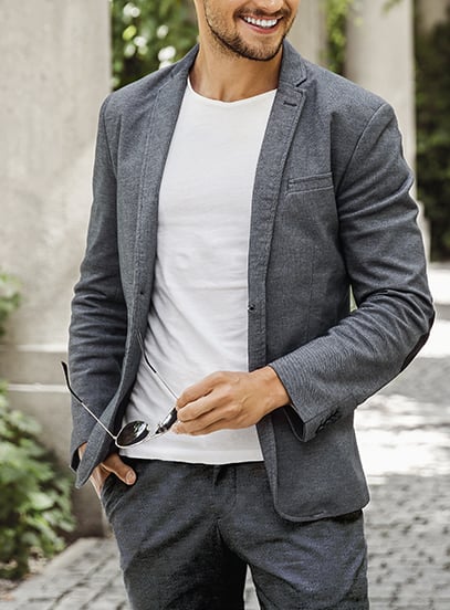 Smiling man in a gray wool-look blazer and white t-shirt holding sunglasses while walking outdoors