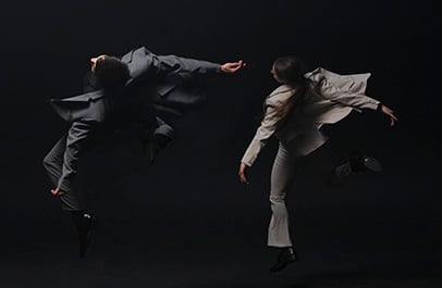 Two dancers in gray and light suits leap mid-air against a dark background