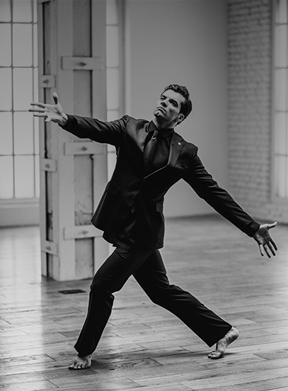 Barefoot man in a dark suit dancing with arms outstretched in a bright room with wooden floor