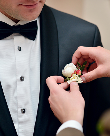 Hands pinning a white rose boutonniere onto the lapel of a man's black tuxedo jacket and bow tie