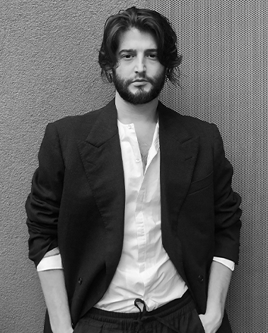 Man with wavy hair and a beard in a dark jacket and white shirt, hands in pockets, against a wall