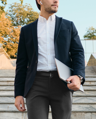 Man in a navy blazer and white shirt walking down stone steps, holding a laptop, wearing a wireless earbud