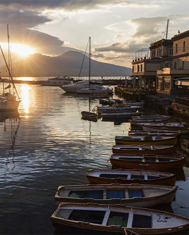 Wooden rowboats moored in a calm harbor at sunset, with sailboats and a mountain in the background