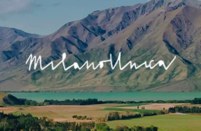 Mountains above a turquoise lake and green fields, with the handwritten text "Milano Unica" overlaid