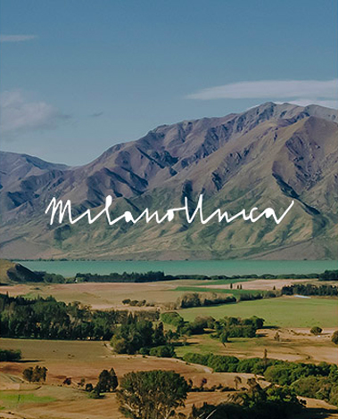 Mountain over a turquoise lake and green farmland, with the handwritten text "Milano Unica"
