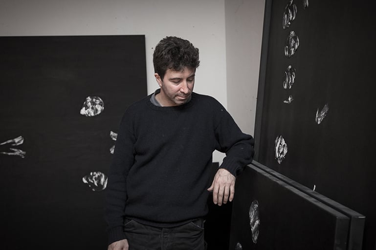Man in a dark sweater leaning on large black canvases with white brushstrokes in a studio