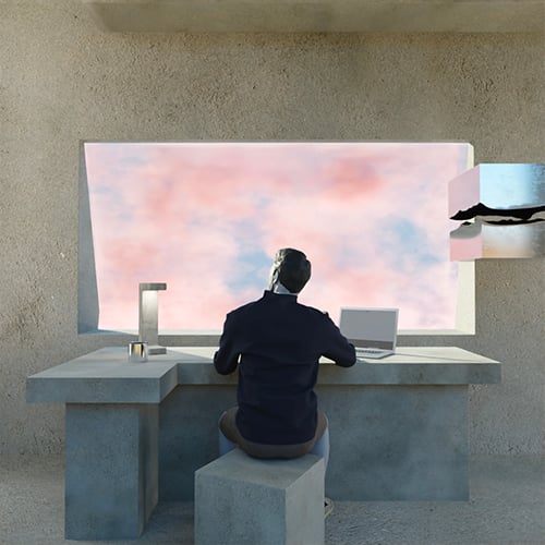 Man in a dark sweater sits at a concrete desk with a laptop, facing a large window onto a pink sky.
