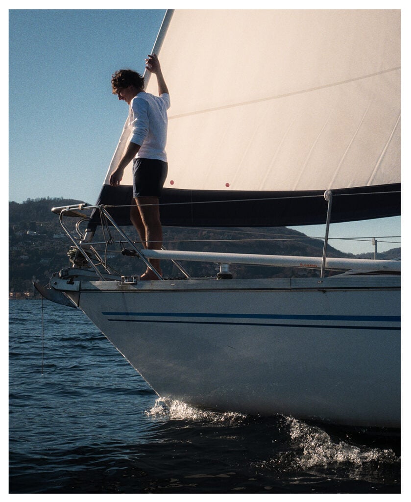 Man in white sweater and navy shorts stands on the bow of a sailboat, holding the sail on a calm lake.