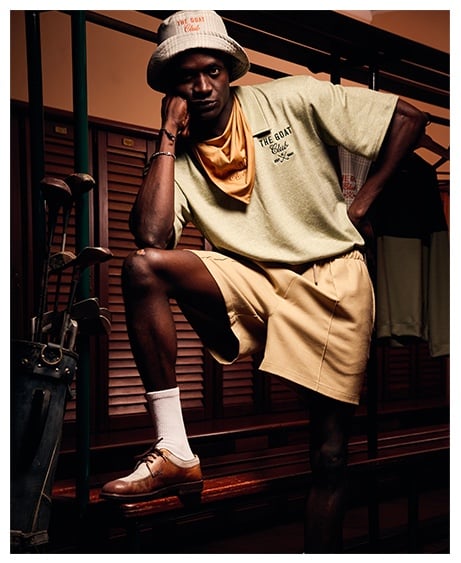 Man in bucket hat, green tee reading "The Goat Club" and beige shorts poses in a locker room beside golf clubs