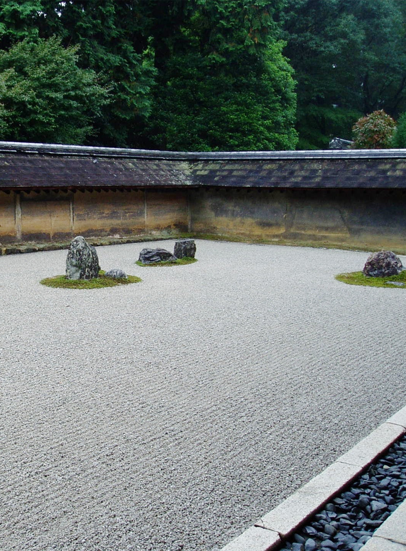 Raked gravel garden with moss-covered rock groups, bordered by a low tiled wall and green trees