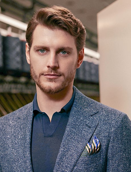 Man with styled hair and a beard wearing a blue textured wool jacket, navy polo and patterned pocket square