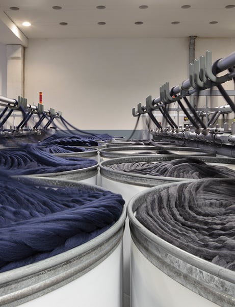 Large white containers filled with blue and grey wool slivers in a textile mill, with overhead machine arms above.
