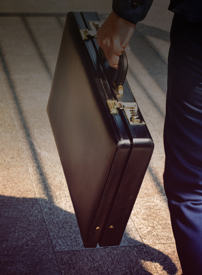 Person in a navy suit carries a black leather briefcase with brass locks across a sunlit stone floor