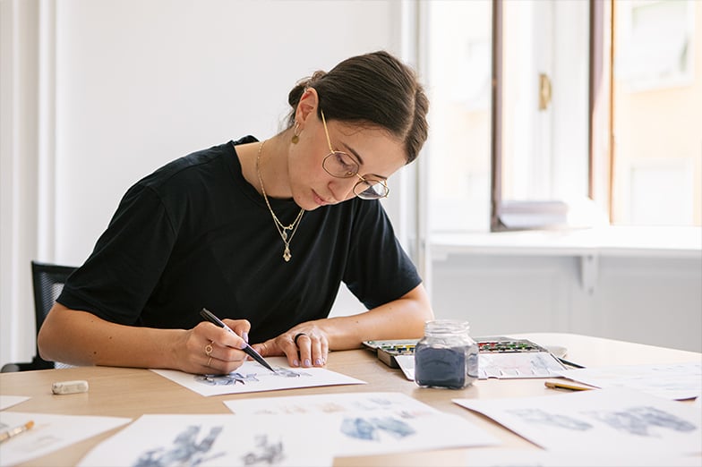 Woman in glasses and a black t-shirt paints with a brush on paper at a table with watercolors and sketches