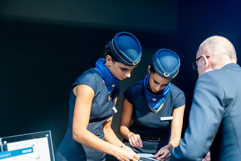 Two flight attendants in blue uniforms and hats look down at a tablet with a man in a suit and glasses