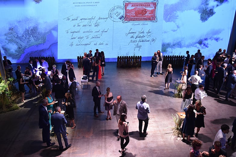 Guests mingle at an evening event in front of a giant projected "Australia Post Card" screen