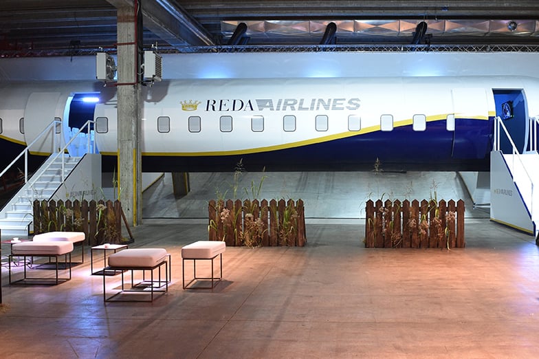 Mock airplane fuselage with Reda Airlines logo in an indoor event space with white ottomans and wooden planters