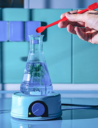 Hand with a red spoon adds material to a flask of liquid on a magnetic stirrer hotplate in a lab