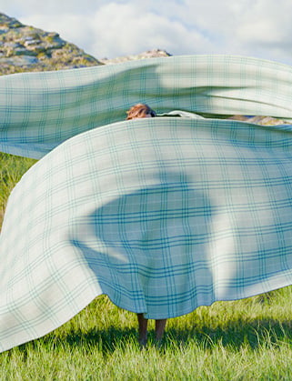 Person hidden behind a large green plaid fabric billowing in the wind on a grassy hillside
