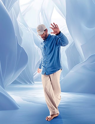 Barefoot man in a blue shirt, beige trousers and cap raises his hand in a pale blue ice cave
