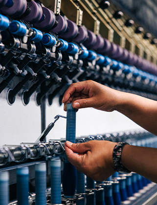 Hands adjusting a blue yarn bobbin on a textile spinning machine with rows of purple and blue spools