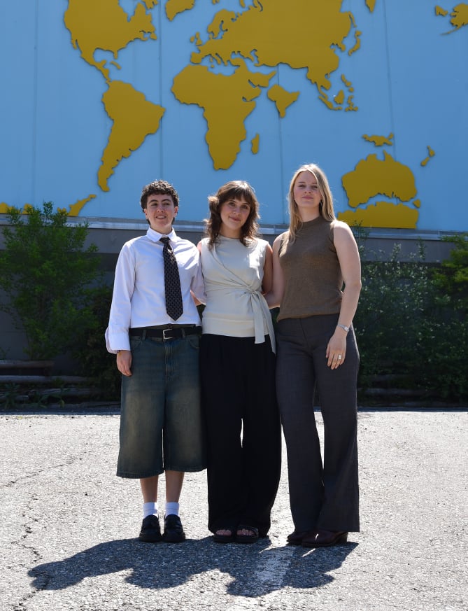 Three people stand together outdoors in front of a blue wall with a yellow world map.